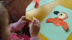 Dwie dziewczynki siedzą przy stoliku i przyklejają do karki kolorowe koła, tworząc papugę techniką płaskie origami z koła.