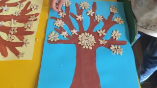 Na stoliku leży praca plastyczne przedstawiająca drzewo namalowane farbami oraz ozdobione nasionami dyni ułożonymi w kształt kwiatów.
