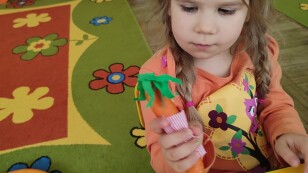 dziewczynka w pomarańczowej bluzce siedzi przy stoliku i prezentuje marchewkę z zielonymi włosami wykonanymi z bibuły oraz w kolorowym stroju z tkaniny w kropki.