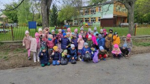 Dzieci ustawione na tle ogrodzenia i przedszkolnego placu zabaw pozują w grupie do zdjęcia po zakończonej akcji sprzątania świata.