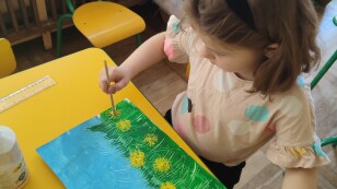 Dziewczynka siedzi przy stoliku i maluje farbami łąkę, żółte kwiaty mniszka i dmuchawce, za pomocą pociętej słomki do napojów.