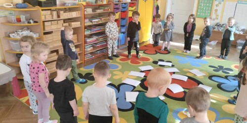 Dzieci stoją w kole na dywanie i uczestniczą w zabawie z kartami z ćwiczeniami
