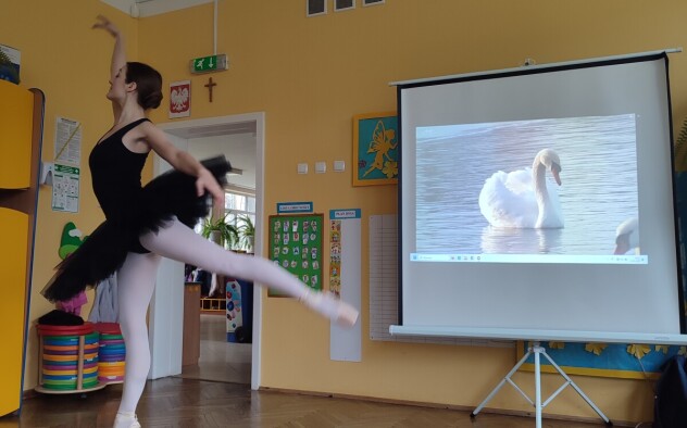 Baletnica wykonuje taniec stojąc na jednej nodze, drugą ma uniesioną do tyłu, w tle widać białego łabędzia