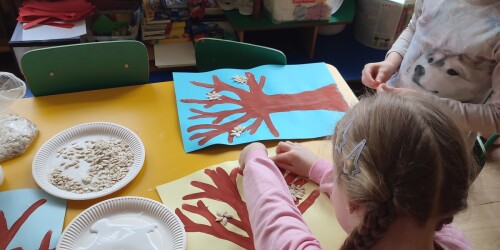 Dwie dziewczynki przyklejają ziarna dyni formując z nich kwiaty na koronie drzewa namalowanego farbami.