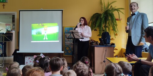 Dzieci siedzą na dywanie przed białym ekranem, na którym wyświetlana jest prezentacja o wiośnie. Obok kobieta śpiewa piosenkę trzymając w dłoniach mikrofon.