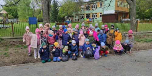 Dzieci ustawione na tle ogrodzenia i przedszkolnego placu zabaw pozują w grupie do zdjęcia po zakończonej akcji sprzątania świata.