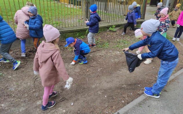 Dzieci poruszają się w okolicy ogrodzenia i szukają śmieci, zbierają je do worków trzymanych przez inne osoby z grupy.