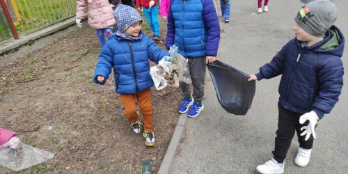 Dwaj chłopcy trzymają worek na śmieci, dookoła nich chodzą dzieci i zbierają pozostawione wokół kawałki folii, papierów, butelki.